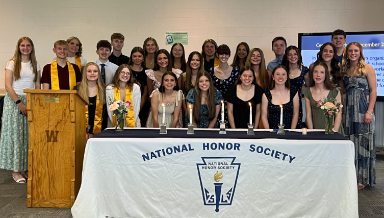 All current members of NHS stand behind a table with 5 candles and two bouquets of flowers. On the left is a wooden podium with a "W" etched on the front.