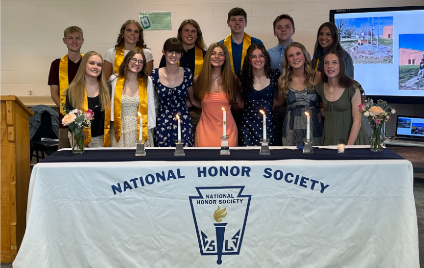 This year's NHS members stand behind a table with 5 candles and two bouquets of flowers. On the left is a wooden podium with a "W" etched on the front.