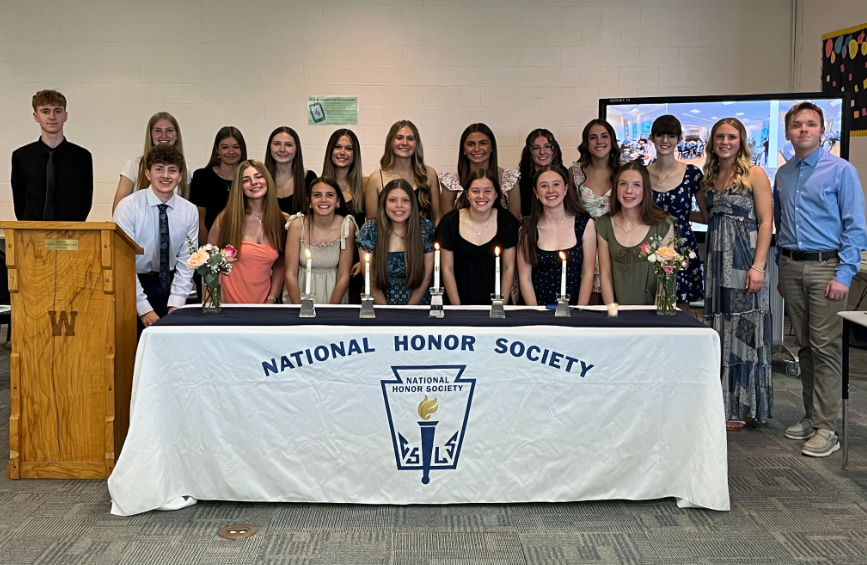 Next year's NHS members stand behind a table with 5 candles and two bouquets of flowers. On the left is a wooden podium with a "W" etched on the front.