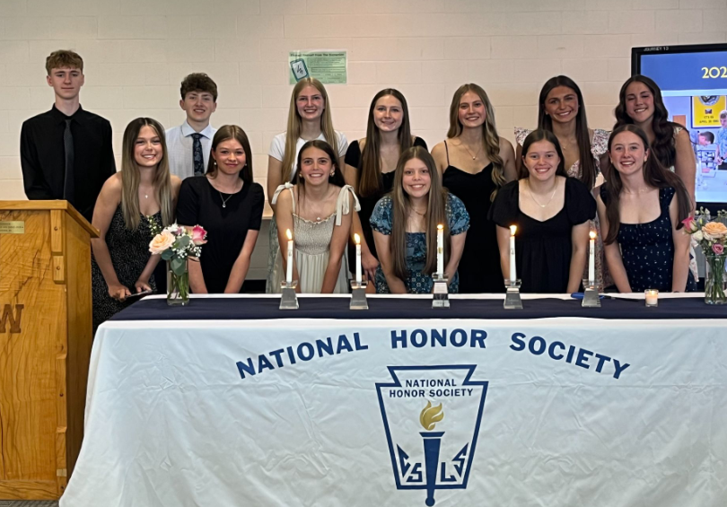 The newest NHS inductees stand behind a table with 5 candles and two bouquets of flowers. On the left is a wooden podium with a "W" etched on the front.