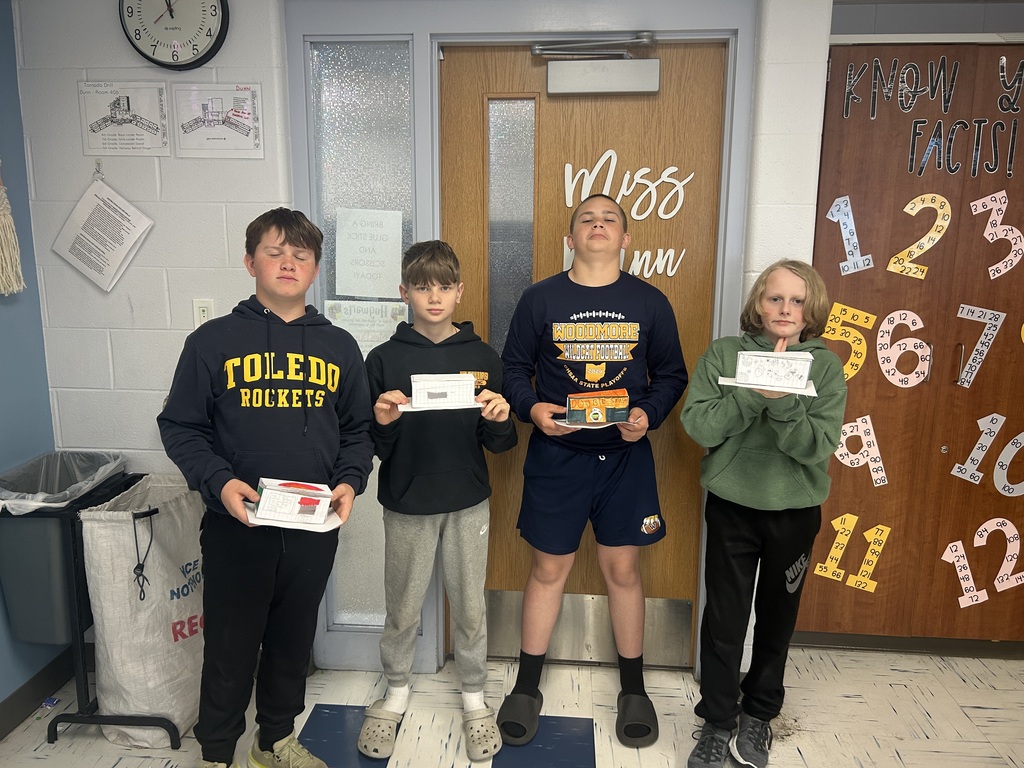 Four students stand by the door of their classroom holding their food trucks in front of them.