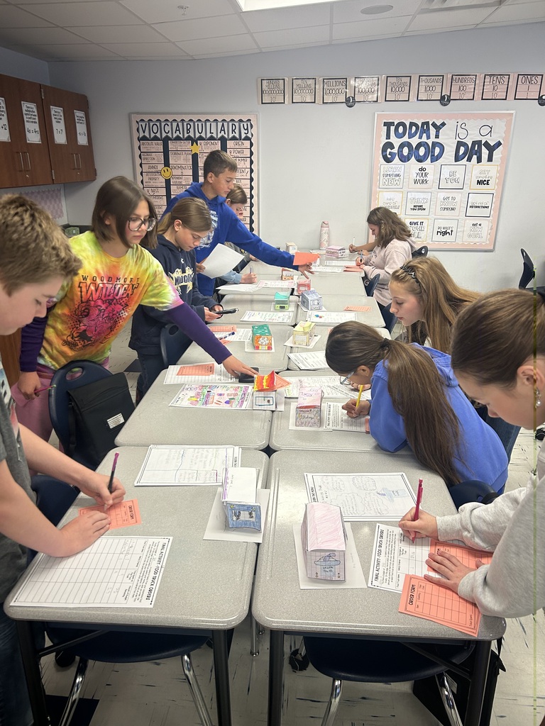 Students are lined up on either side of a row of desks looking at food trucks that are lined up and writing down information.