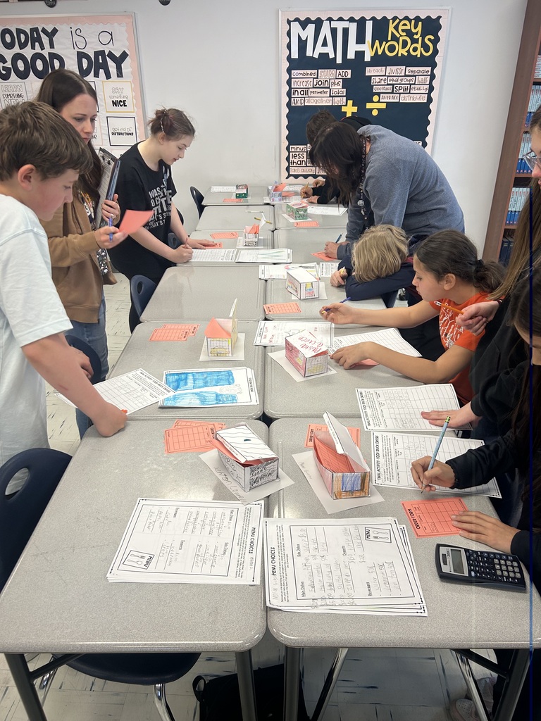 Students are lined up on either side of a row of desks looking at food trucks that are lined up and writing down information.