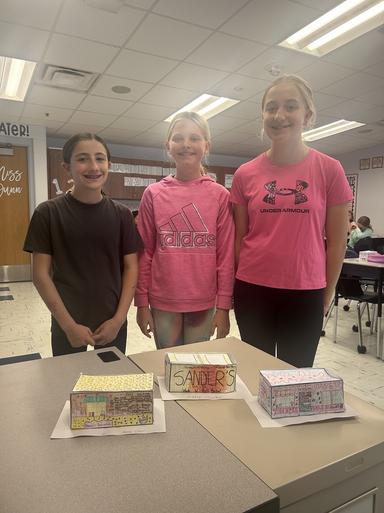 Three students stand behind a desk, smiling for the camera. In front of them, on the desk, are their three food trucks.