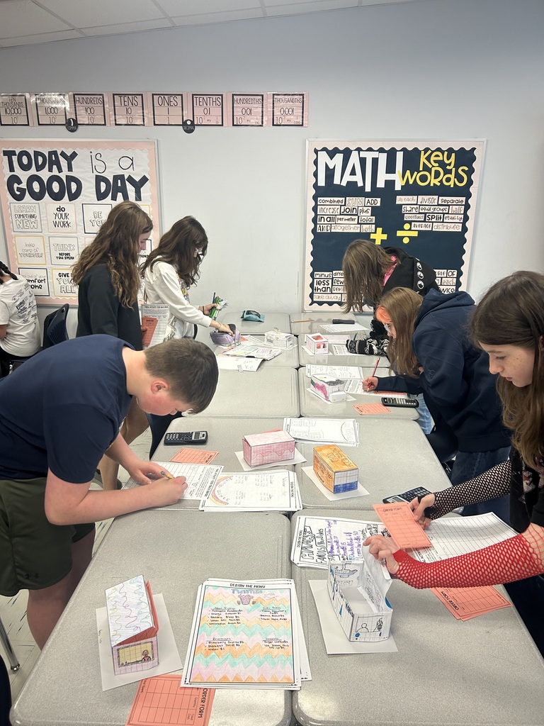 Students are lined up on either side of a row of desks looking at food trucks that are lined up and writing down information.