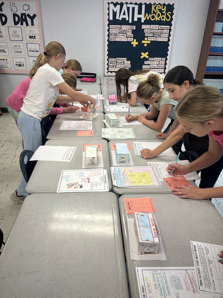 Students are lined up on either side of a row of desks looking at food trucks that are lined up and writing down information.