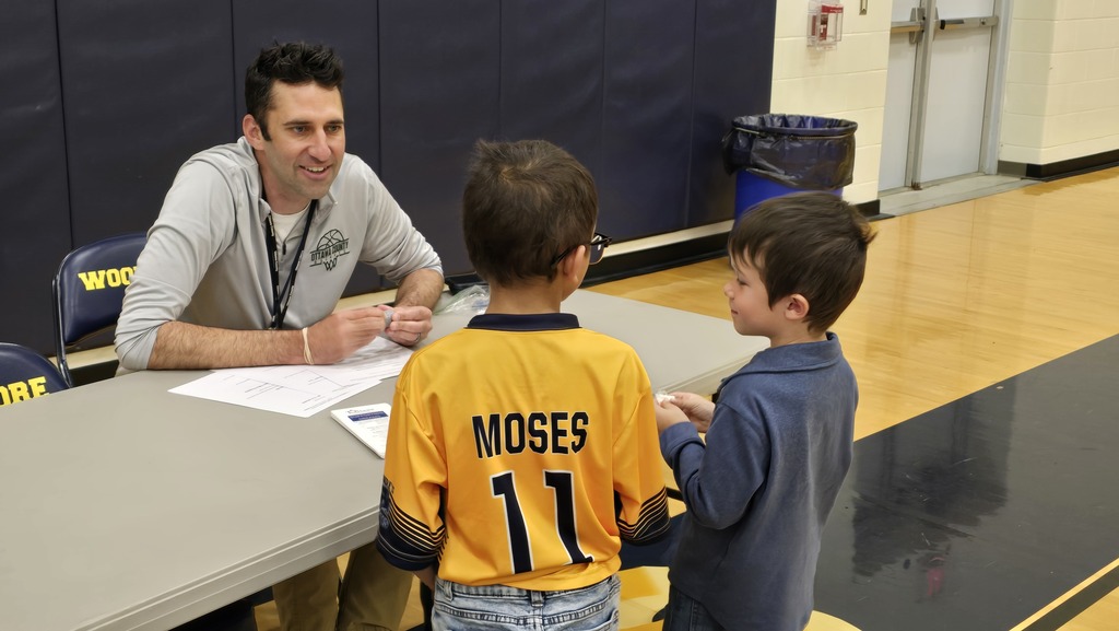 An adult sits at a table with two students standing on the other side, speaking about his career.