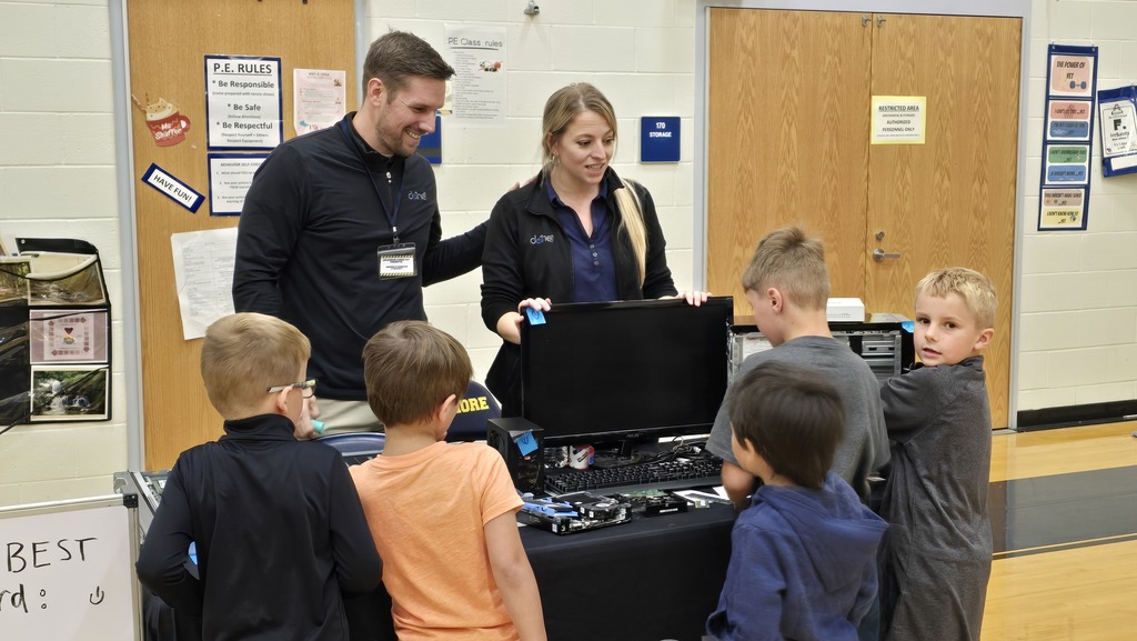 Two adults stand behind a table covered in computer parts in various states of disassembly. Five students stand on the other side of the table looking at all of the parts and listening to the adults speak about their careers.