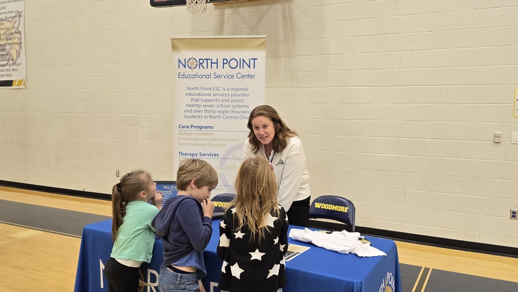 Students stand at a table, listening to a North Point Educational Service Center employee speak about her career. There is an informational banner behind her.