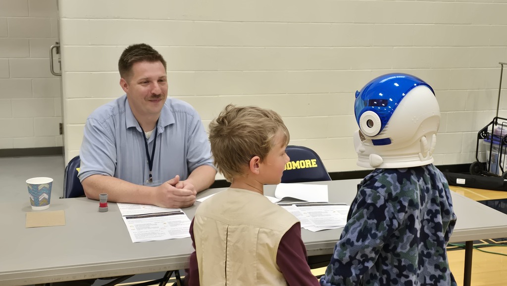 An adult sits at a table with two students standing on the other side, speaking about his career.