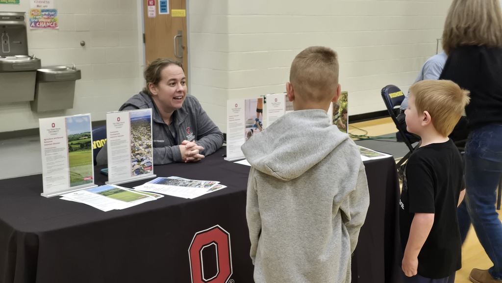 An employee of Ohio State sits at a table covered in a black OSU tablecloth with informational papers sitting on the table. Students stand in front of her asking questions.