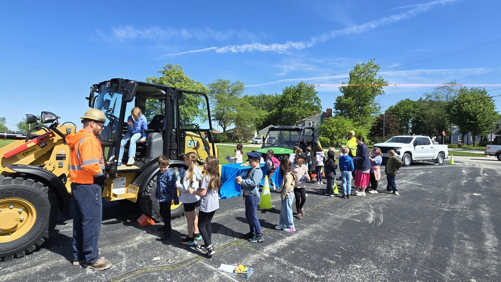 Students line up next to a piece of heavy machinery and await their turn to get inside the cab. It is a bright sunny day with blue skies. An adult stands by the door to the cab of the vehicle discussing his career with the waiting students.