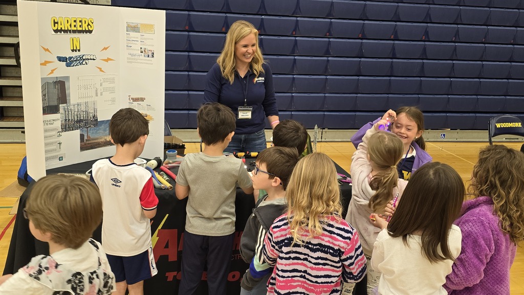An adult stands behind a table with a tri-fold poster board set on a table. A group of students stands on the other side of the table while she explains her career.