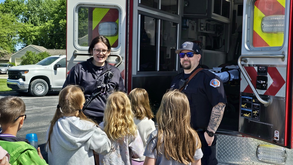 Two adults are at the back of an open ambulance speaking with students about their career.