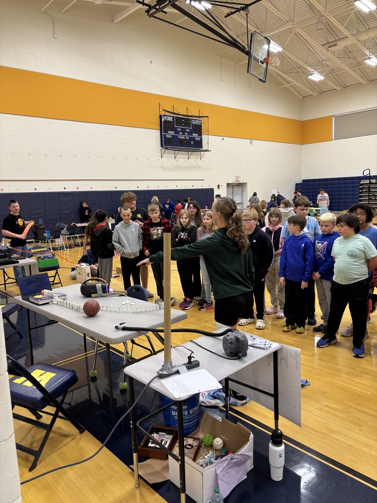 Students gather around a Rube Goldberg machine in the gym as the creators explain and demonstrate their machine.
