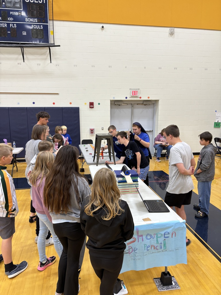 Students gather around a Rube Goldberg machine in the gym as the creators explain and demonstrate their machine.