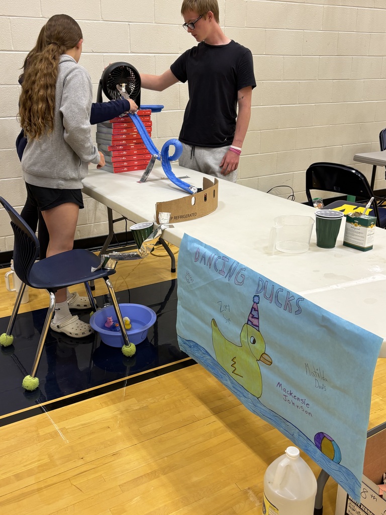 Three students work on setting up their Rube Goldberg machine on a folding table.