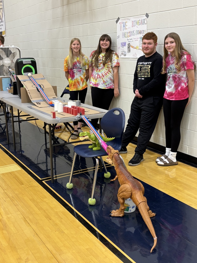 Four students stand against the gym wall with their Rube Goldberg machine in front of them.