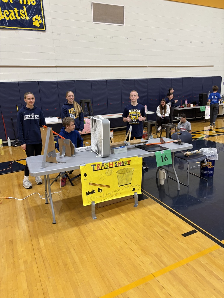 Four students stand behind their Rube Goldberg machine, setup on a folding table.