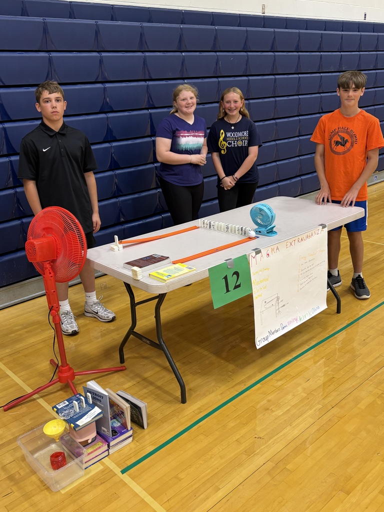 Four students stand behind their Rube Goldberg machine, setup on a folding table.