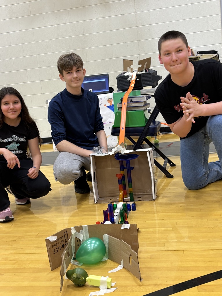 Three students kneel on the gym floor next to their Rube Goldberg machine.