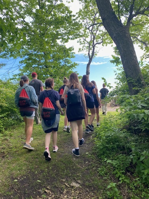 Students are hiking on a nature trail around Gibraltar Island. They are in the middle of trees with blue skies in the background.