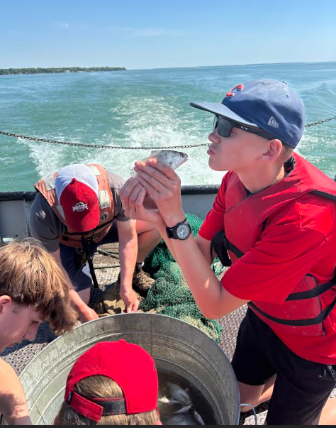 Students are on board a boat (you can see wake in the background). A student holds up a fish, pretending to kiss it.