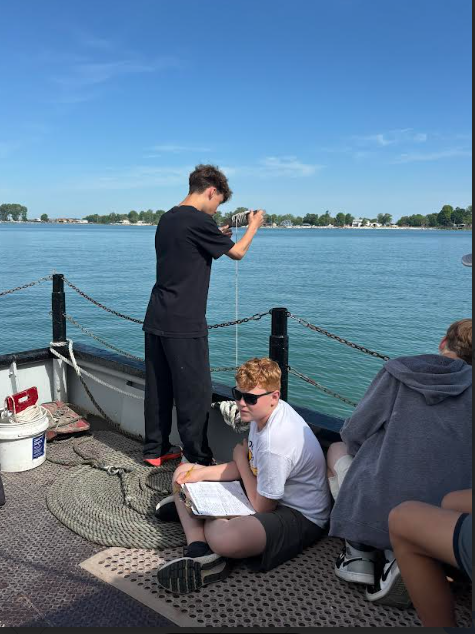 A student stands by the water, dropping a rope into the water to take a measurement. Another student sits nearby, clipboard in hand to write the measurement down.