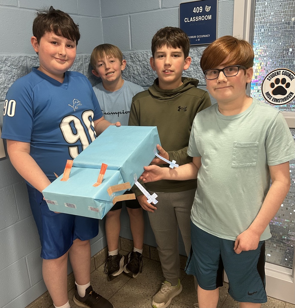 Four students hold their zoo shaped like an axolotl in front of their classroom door.