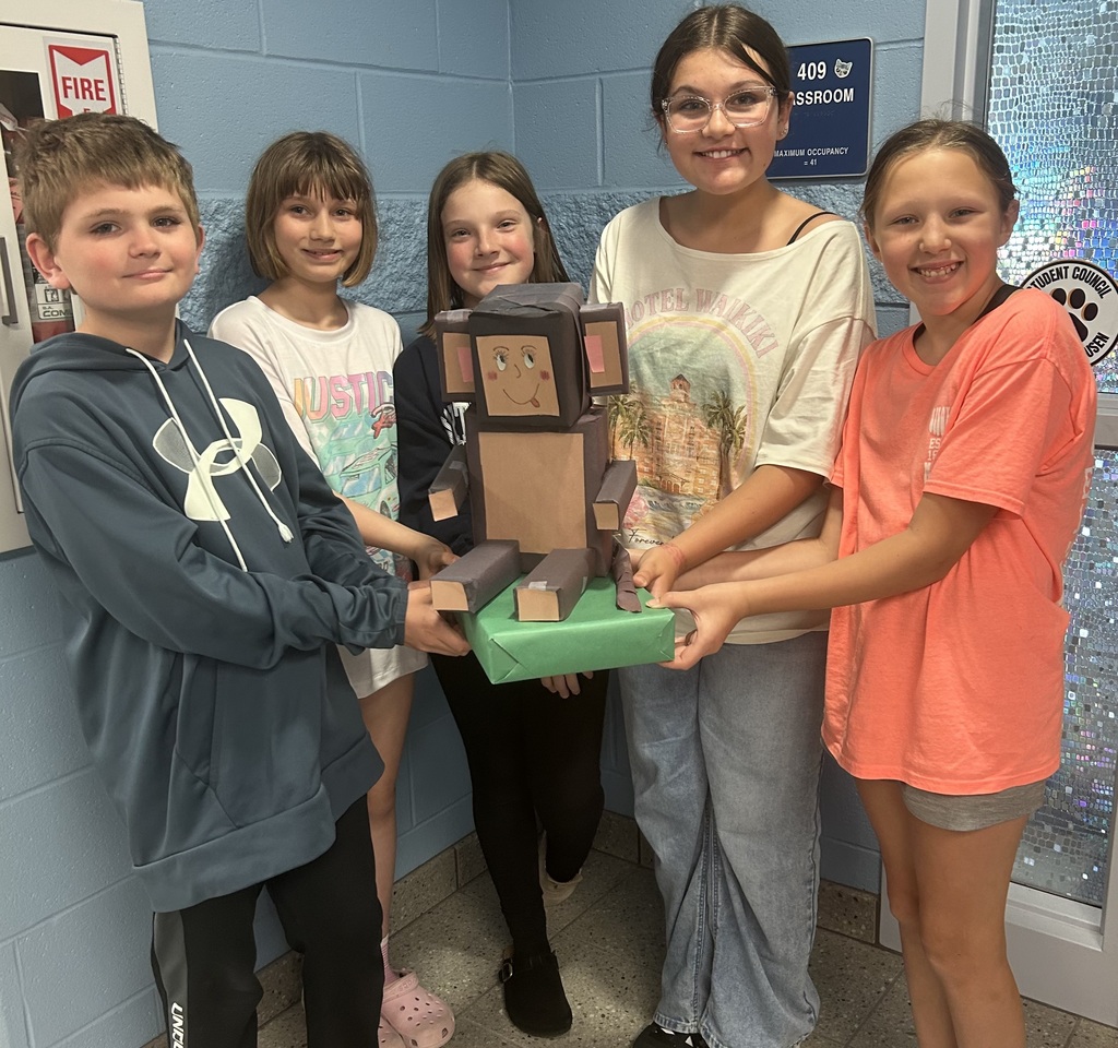 Five students hold their zoo shaped like a monkey, posing in front of their classroom door.