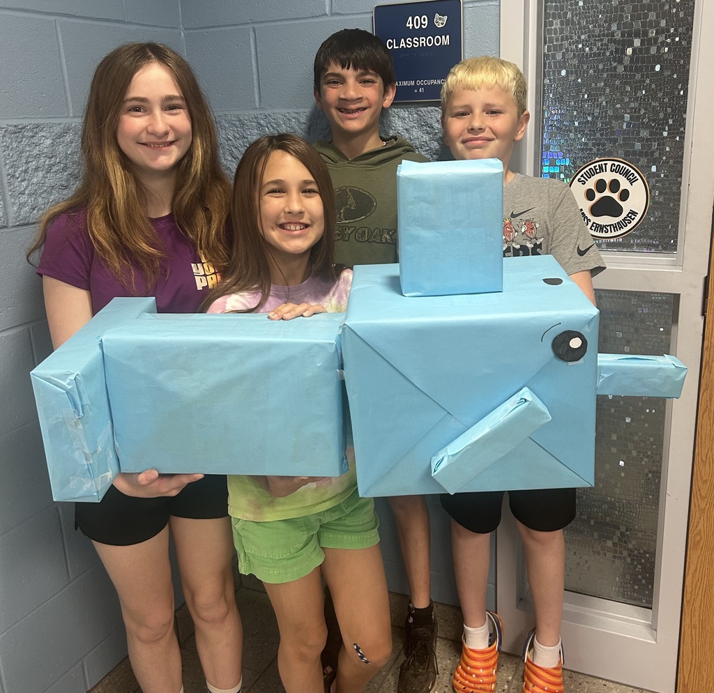 Four students hold their zoo shaped like a narwhal in front of their classroom door.