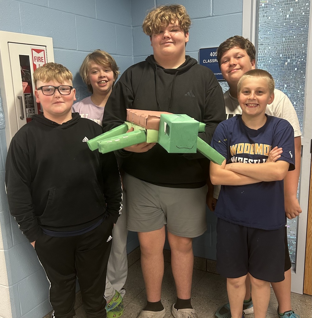 Five students hold their zoo shaped like a turtle while standing in front of their classroom door.