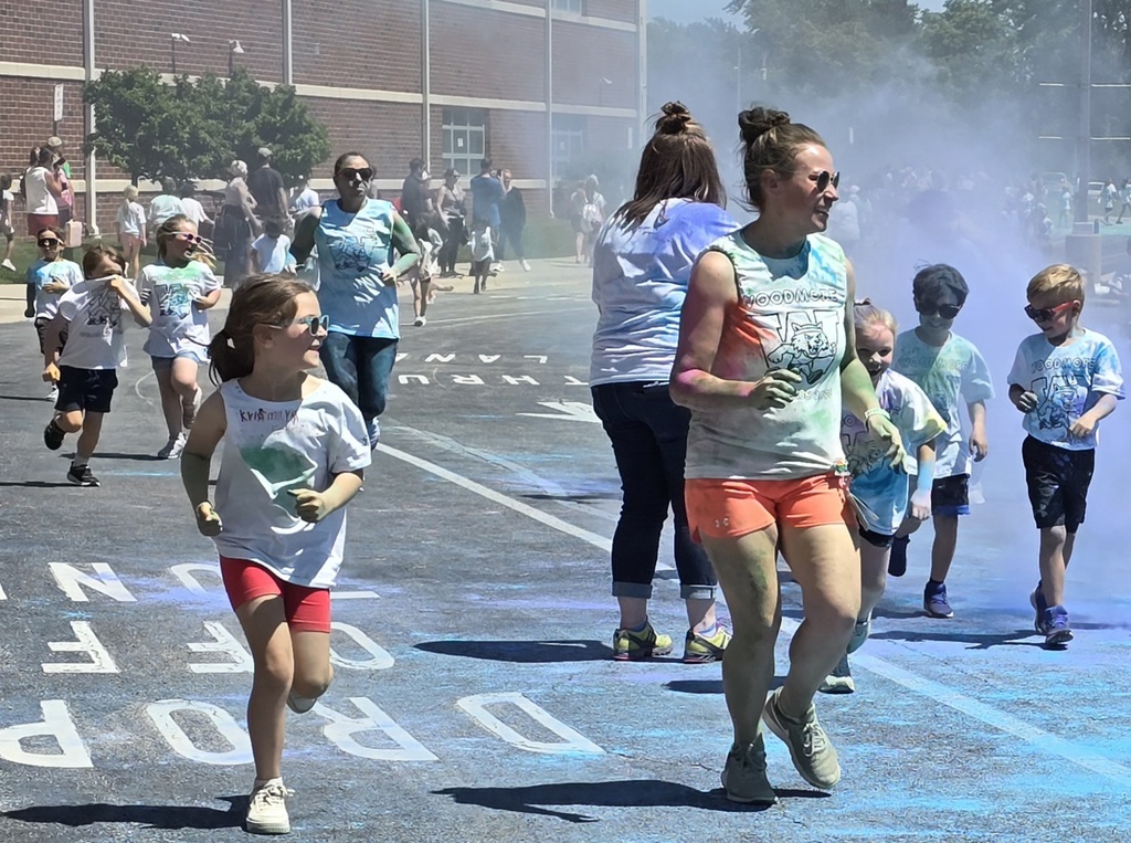 Students and kindergarten teachers are running out in front of the school covered in colorful powder.