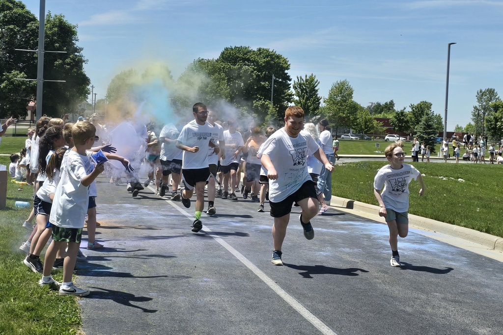 Students run through clouds of colored powder out front of the school on a day with lots of sunshine and bright blue skies. Other students stand to the side, throwing the powder.