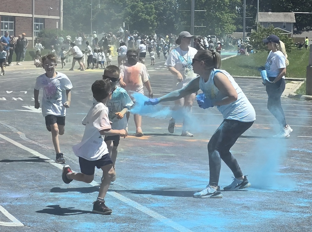 Students covered in colorful powder run outside of the school and get blue powder tossed at them by a teacher.