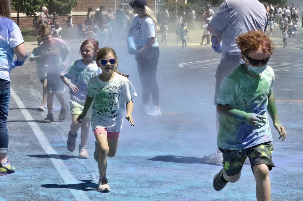 Students run through teachers tossing blue powder. Some wear sunglasses, one has on a face mask.