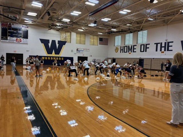 Students play a game on the gym floor using pom poms. Other students are onlookers.