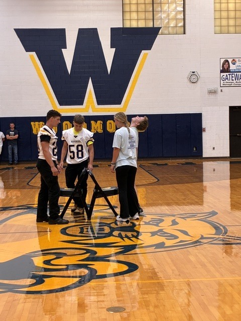 Students are playing a game in the middle of the gym floor. One student has his head thrown back in exasperation. They are all circling chairs on the floor.