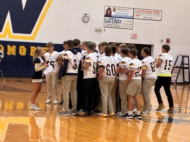 A cheerleader speaks with a group of football players grouped on the gym floor.