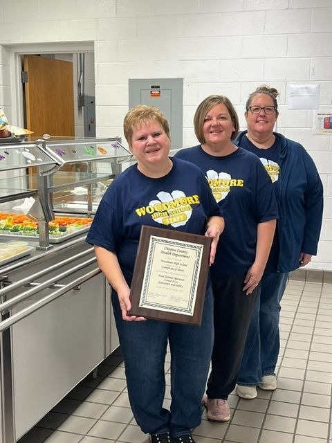 Three Woodmore High School cafeteria staff members wearing matching navy blue "Woodmore High School Staff" t-shirts stand in a cafeteria setting. The woman in the front proudly holds up the framed Certificate of Merit from the Ottawa County Health Department, with food service counters visible behind them.