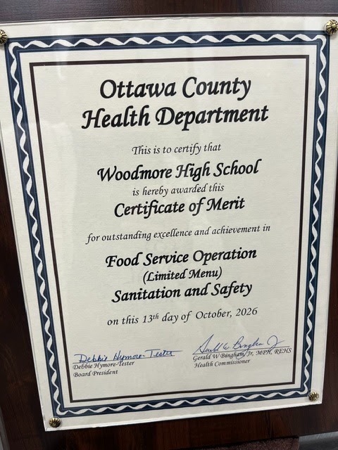 A framed Certificate of Merit awarded by the Ottawa County Health Department to Woodmore High School on October 13, 2026. The certificate recognizes outstanding excellence and achievement in Food Service Operation (Limited Menu) Sanitation and Safety, signed by Board President Debbie Hymore-Tester and Health Commissioner Gerald W. Bingham Jr