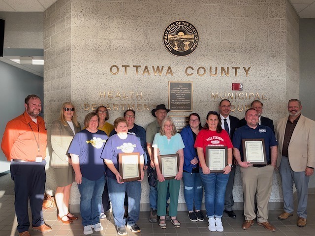 A group photo of various recipients and officials gathered in front of an Ottawa County Health Department building wall displaying the Great Seal of the State of Ohio. Several people hold framed award certificates, including the Woodmore High School staff members.