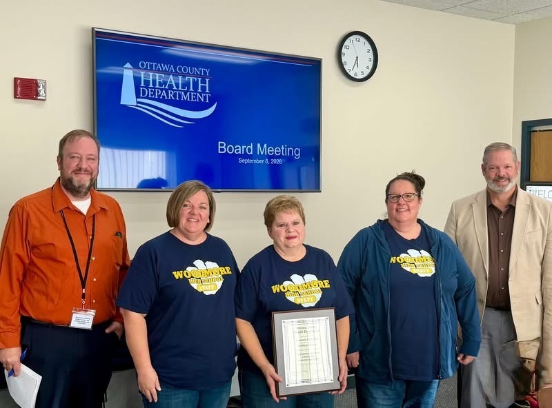 Three Woodmore High School staff members stand with two officials inside a room in front of a digital display monitor that reads "Ottawa County Health Department Board Meeting". The staff member in the center holds the framed Certificate of Merit.