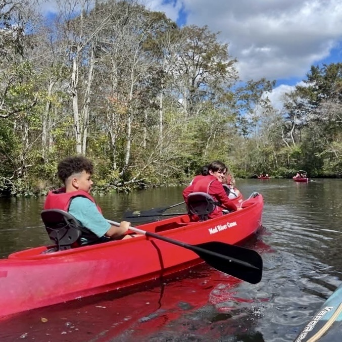 Students paddle red canoes along a calm river surrounded by trees.