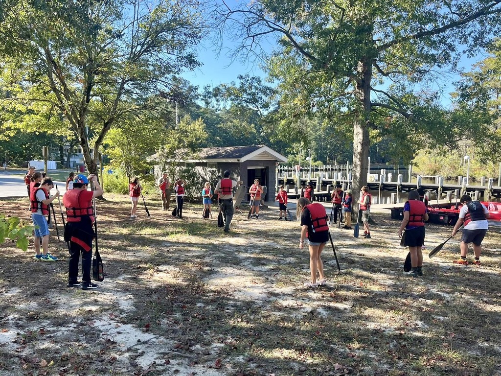 A large group of students in life jackets practice canoe paddling techniques on shore before heading into the water.