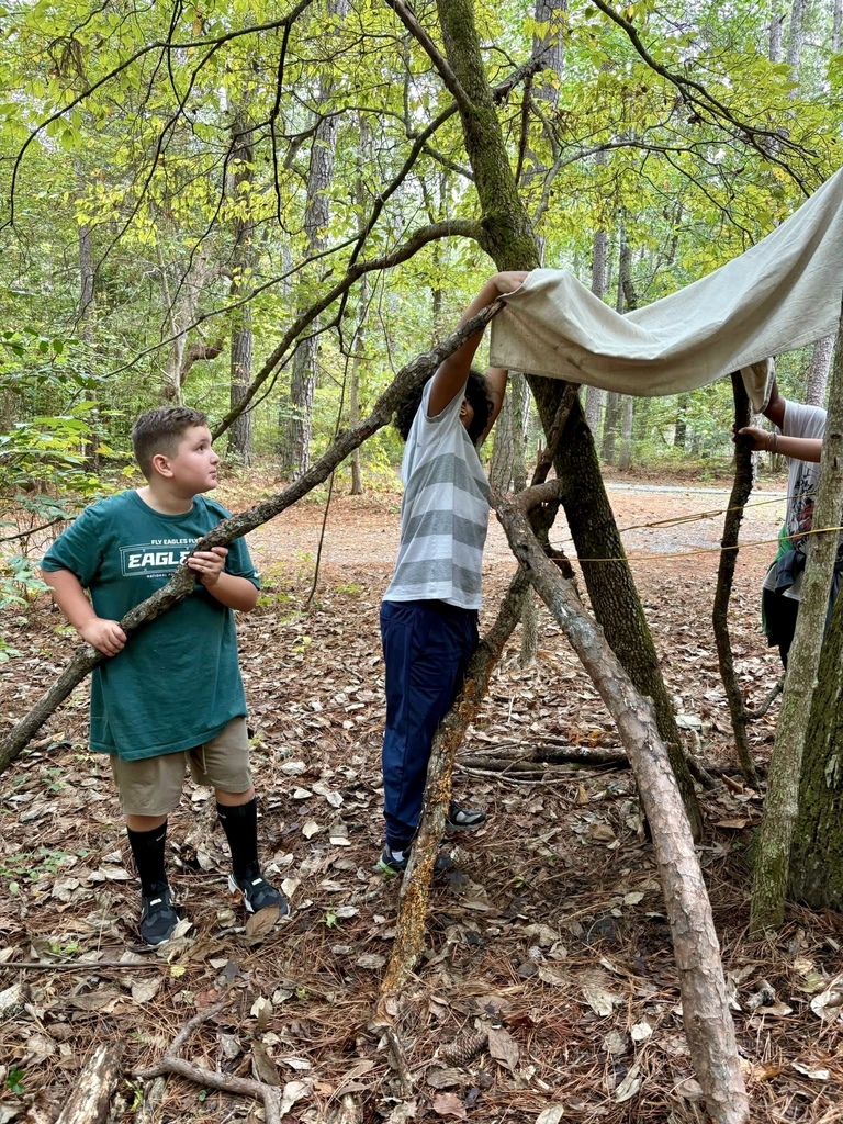 Two students work together to build a shelter in the forest using branches and a tarp.