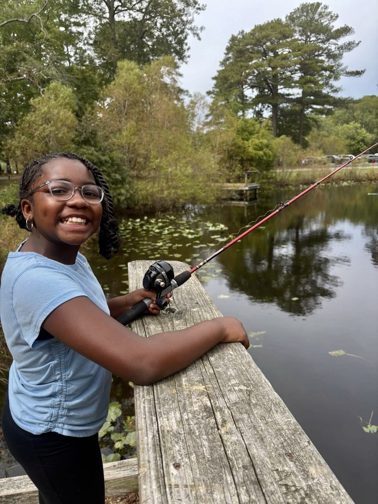 A student smiles while holding a fishing rod on the edge of a pond.