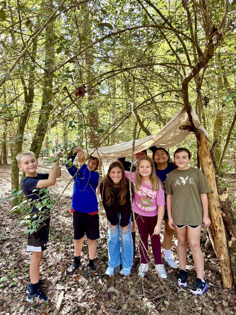 A group of students stand proudly by a completed forest shelter made of branches and a tarp.
