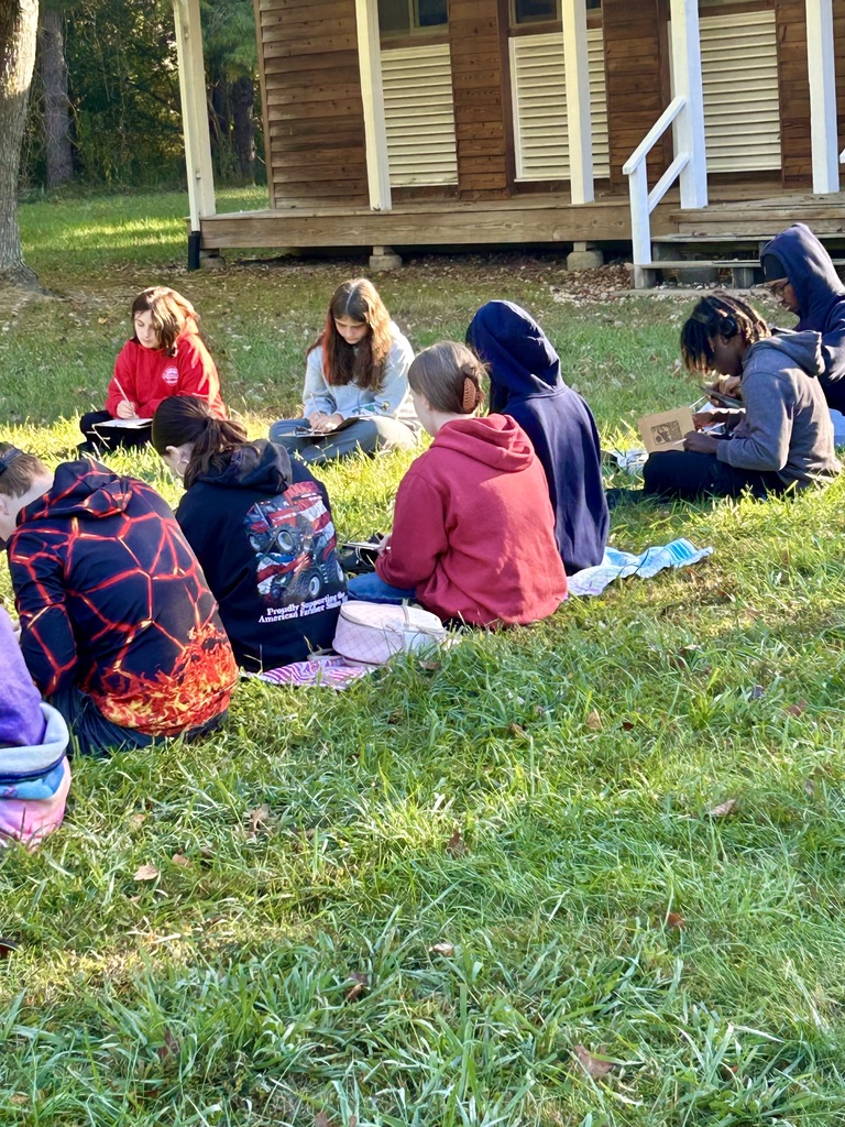 Students sitting on the grass outside a cabin, writing in journals.