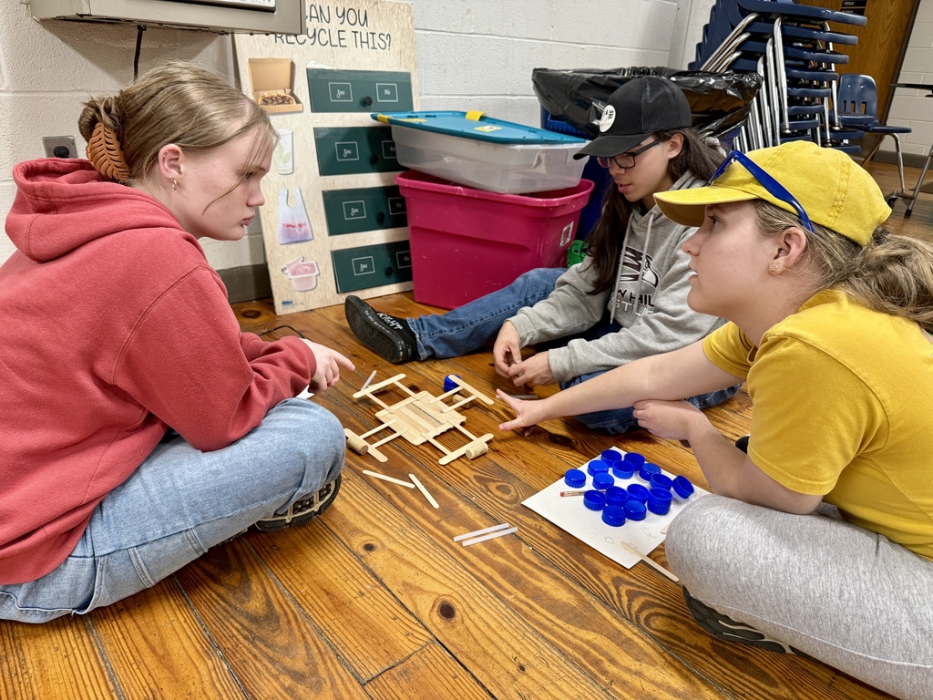 Three students indoors building a small structure out of craft sticks and bottle caps.